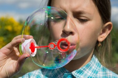Bir kız bir ekose gömlekli sabun köpüğü ile oynuyor. Kızın gözleri kapalı. Arkasındaki kız yeşil çim ve sarı çiçekleri vardır.
