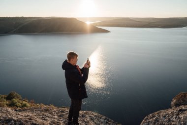 Yan görünüm genç adam güneşli nehir kıyısında duran ve smartphone ile fotoğraf çekme
