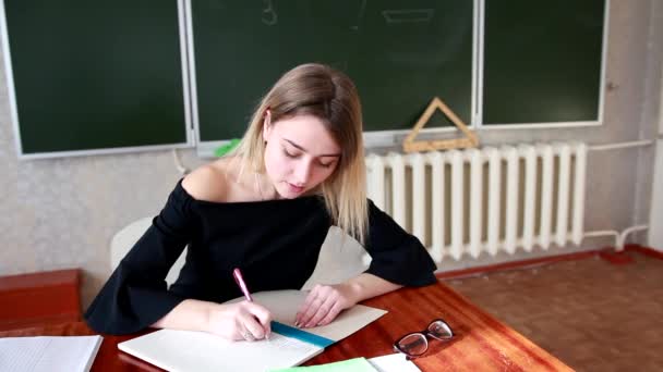 一位严格的年轻金发老师在学生的笔记本上检查作业 她对工作结果不满意 黑板上的铭文 在课堂上工作 — 图库视频影像
