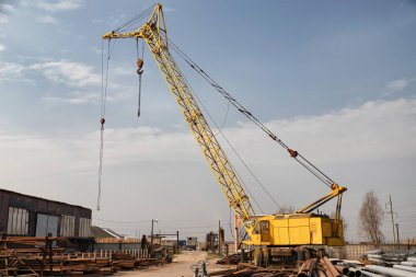Büyük sarı bir vinç, metal borular, montajlar ve metal yapılar arasında yuvarlanan metalin temelinde yer alır.