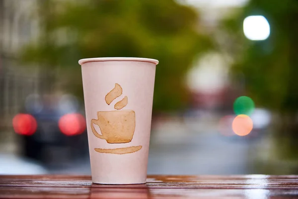 White paper cup with a picture of a cup stands on a wooden table in the background of a blurred street
