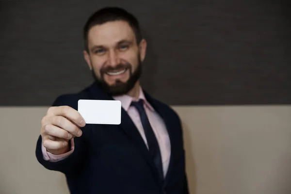 A white empty card in the hands of a blurred bearded young man with a smile in a business suit.