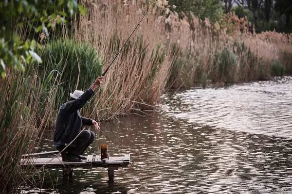 A man sits in the rain on the bridge and catches fish in the pond.
