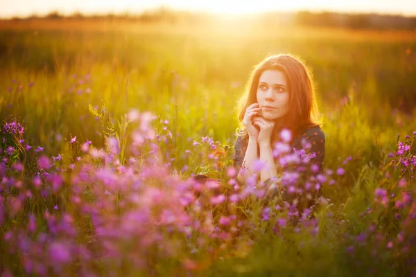 A young adult European girl with long fair-haired hair sits on a flowering meadow, holding her hands in front of her face. Pink flowers in the foreground. Beautiful morning light.