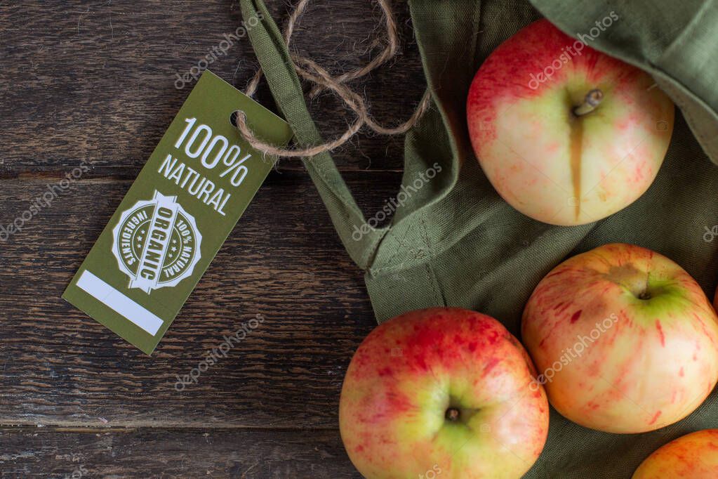 Juicy red apples and a green bag of cotton fabric with a label 100 natural product, lie on a textured table made of dark brown wood