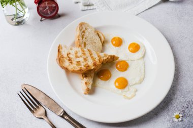 Kahvaltı vakti geldi. Kahvaltıda kızarmış yumurtalı tost, açık gri bir arka plan, gazete ve çalar saat.