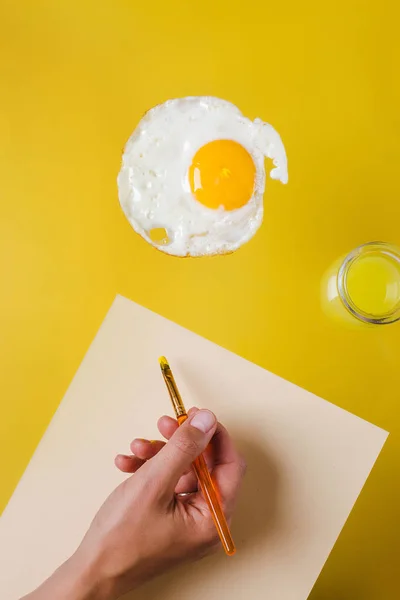 La main avec un pinceau et une feuille de papier blanc dessine des œufs frits, à côté d'un pot d'eau jaune — Photo