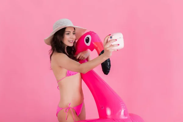 Девушка в купальнике и шляпе держит фламинго и делает селфи фото на мгновенной камере — стоковое фото