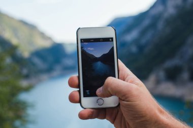 Bir adam telefonla dağ manzarasının fotoğrafını çekiyor. Bir elinde tutuyor.
