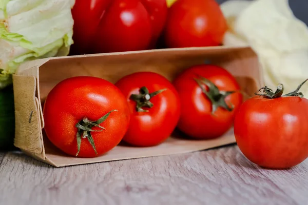 Fresh tomatoes in a box on a light countertop on a background of cabbage and lettuce. Still life, composition of fresh vegetables.