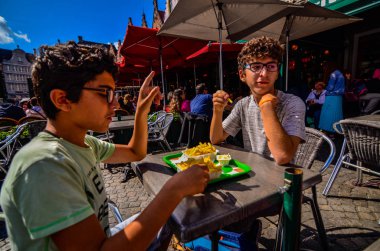 Bruges, Belçika, Ağustos 2019. Turistik Markt Meydanı 'na bakan bir kızartma dükkanı. İki beyaz çocuk mayonezli cipsin tadını çıkarıyor..