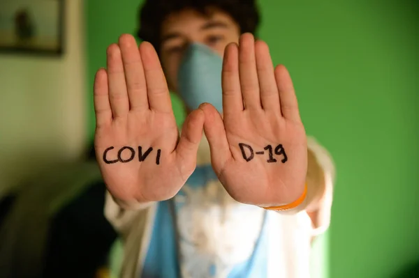 Close-up image of the palms of the hands with the inscription COVID-19 of a Caucasian boy with curly brown hair wearing a blue mask. Conceptual image of danger of contagion. Selective focus on palms.
