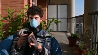 Terasta güneşlenirken koruyucu maske takan beyaz bir çocuğun portresi akıllı telefonuyla meşgul. Sağlık ve güvenlik yaşam konsepti, virüs koruması, salgın.
