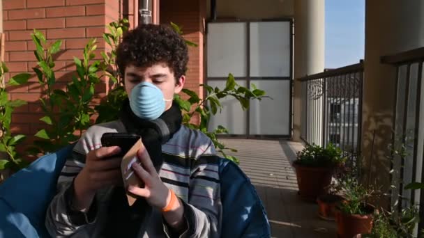 Porträtt av en ung kaukasisk pojke bär en skyddande mask medan du njuter av lite sol sitter på terrassen, är upptagen med sin smartphone. Koncept för hälsa och säkerhet, virusskydd, pandemi. — Stockvideo