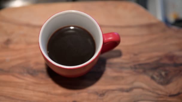 Close-up afbeelding van een rode koffiekop aan de buitenkant en wit aan de binnenkant rustend op een houten snijplank. Er komt een druppel stoom uit de beker. wazige achtergrond. — Stockvideo