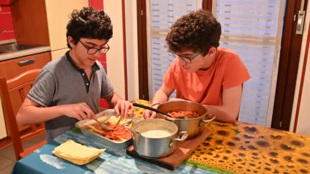 Hausgemachte Lasagne zubereiten. Zwei kaukasische Jungen gießen die Fleischsoße über den Blätterteig und streichen ihn mit Löffeln. — Stockvideo