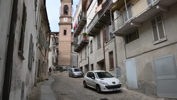 Costigliole d 'Asti, Piedmont, İtalya. Mart 2020. Kırmızı tuğlalı çan kulesinde eğimli bir görüntü. Caddedeki evlerin perspektifi çan kulesine bakıyor. İki araba park edilmiş. — Stok video