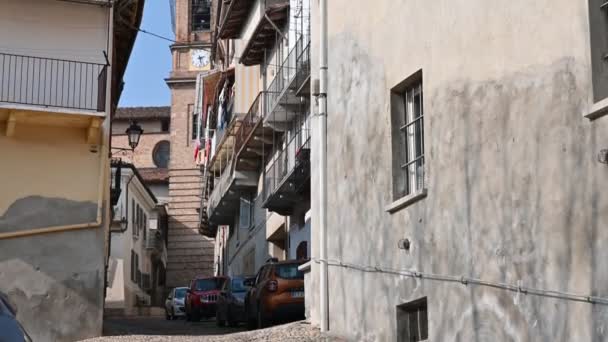 Costigliole d'Asti, Piémont, Italie. Mars 2020. Images avec inclinaison sur le clocher en brique rouge. La perspective des maisons le long de la rue amène le regard vers le clocher. Deux voitures garées — Video