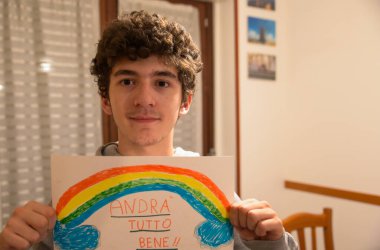 Turin, Piedmont, Italy. March 2020. Cornonavirus quarantine. Portrait of a brown-haired caucasian curly-haired boy holding up a drawing with the rainbow and the message will be all right! clipart
