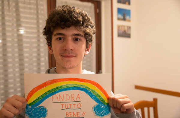 Turin, Piedmont, Italy. March 2020. Cornonavirus quarantine. Portrait of a brown-haired caucasian curly-haired boy holding up a drawing with the rainbow and the message will be all right!