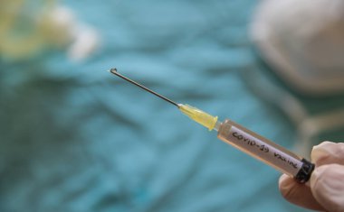 Coronavirus aşısı olan bir şırınganın yakın çekim görüntüsü. Onu tutan el pompaya basıyor ve iğneden bir damla çıkıyor. Bulanık arka planda bir maske ve dezenfekte edici jel var.