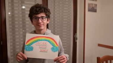 Torino, Piedmont, İtalya. Mart 2020. Cornonavirus karantinası. Kahverengi kıvırcık saçlı ve gözlüklü beyaz bir çocuğun portresi elinde gökkuşağıyla bir çizim ve mesaj iyi olacaktır.!