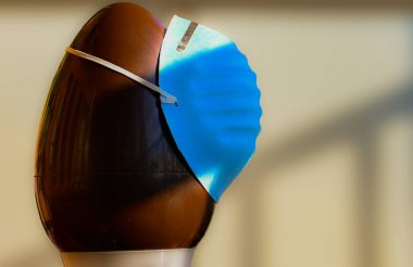 Conceptual close-up image of Easter at the time of the coronavirus pandemic. A chocolate egg, with an olive branch next to it, wears a blue protective mask on the egg. clipart