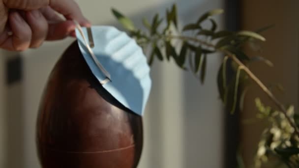 Conceptual close-up image of Easter at the time of the coronavirus pandemic. A chocolate egg with an olive branch next to it. Caucasian man hands put a blue protective mask on the egg. — Stock Video