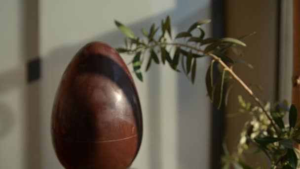Fogalmi közeli kép húsvétról a koronavírus világjárvány idején. Egy csokitojás olajággal mellette. Fehér férfi keze kék védőmaszkot tett a tojásra.. — Stock videók