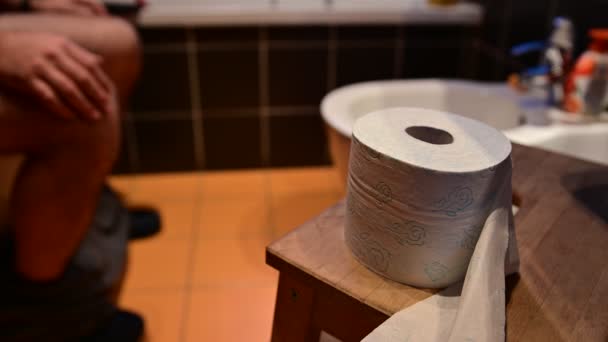 Close-up image of the toilet paper.In the blurred background you can recognize the bare legs of a Caucasian man as he defecates.The hands drum the fingers.The right hand tries to grab the toilet paper — Stock Video