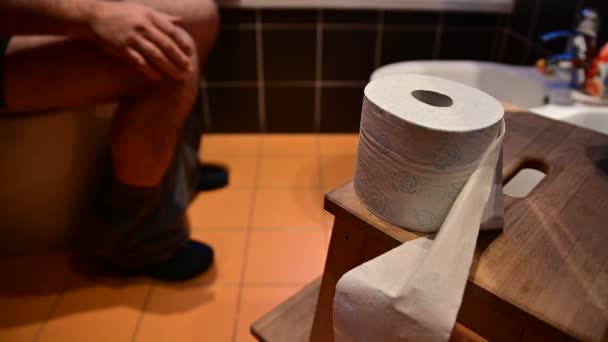 Close-up afbeelding van het toiletpapier.In de wazige achtergrond herken je de blote benen van een blanke man als hij ontlasting heeft.De handen trommelen de vingers.De rechterhand probeert het toiletpapier te grijpen — Stockvideo