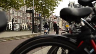 Amsterdam, Hollanda - Ağustos 2019: Bulanık bir çerçeveden ve park halindeki bir bisikletin eyerinden bisikletçilerin bisiklet yolundan geçtiğini görüyoruz. Biraz daha sola, tramvay durağına.. 
