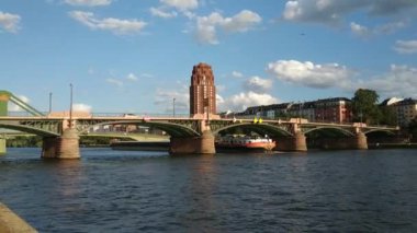 Frankfurt, Almanya - Ağustos 2019: Ana Köprü 'nün altından bir mavna geçiyor. Beyaz bulutlu mavi gökyüzü. Sağdaki ana plaza binası. İnsanlar nehirde yürüyor. pan 30fps.