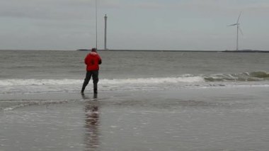Hollanda, Zealand bölgesi - Ağustos 2019: Rüzgar türbinleri kuzey denizi kıyısındaki kumsalın arka planıdır. Kırmızı ceketli bir balıkçı..