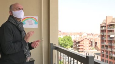 Kafkasyalı adam beyaz bir maske takıyor, balkondan yakındaki evlere bakıyor ve koronavirüs için ani kalabalığı alkışlıyor. Duvardaki çizimde gökkuşağı var. Torino, Piedmont, İtalya