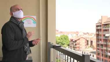 Kafkasyalı adam beyaz bir maske takıyor, balkondan yakındaki evlere bakıyor ve koronavirüs için ani kalabalığı alkışlıyor. Duvardaki çizimde gökkuşağı var. Torino, Piedmont, İtalya