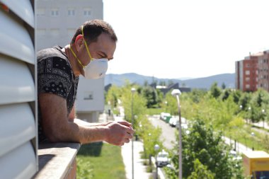 Maskeli orta yaşlı bir adam evde kalıyor, balkondan bakıyor. 