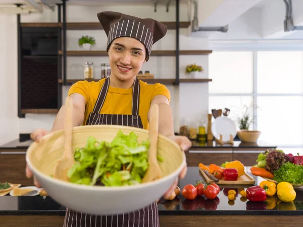 The housewife dressed in an apron and a hair cap, smiled and offered a salad bowl on the front. Morning atmosphere in a modern kitchen. The kitchen counter full of various kinds of vegetables.