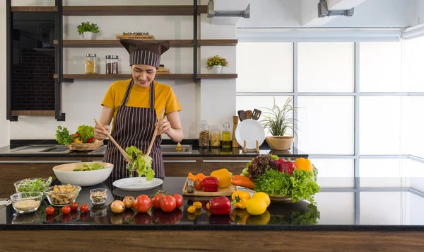The housewife dressed in an apron and a hair cap, mix the vegetables in a salad bowl together with a wooden ladle. Morning atmosphere in a modern kitchen.