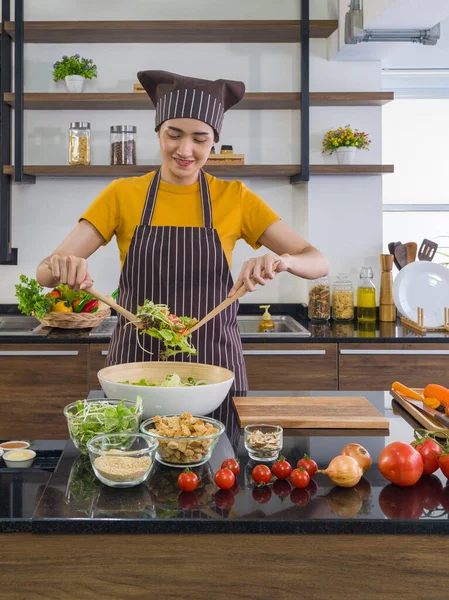 The housewife dressed in an apron and a hair cap, mix the vegetables in a salad bowl together with a wooden ladle. Morning atmosphere in a modern kitchen.