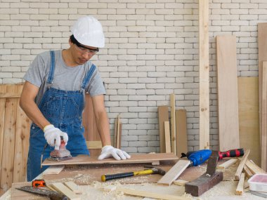 Sert şapkalı ve toz geçirmez gözlüklü Asyalı marangoz zımpara kağıdıyla aşınmış kontrplak yüzeylerini pürüzsüz hale getirmek için zımpara makinesi kullanıyor. Atölyede sabah çalışma ortamı.