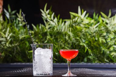 Bir küçük düşük bir kırmızı alkollü kokteyl ile büyük kare parça güzel bulanık yeşil bitkilerin bir arka plan açık buz dolu bir cam vazo yanındaki masada camdır.