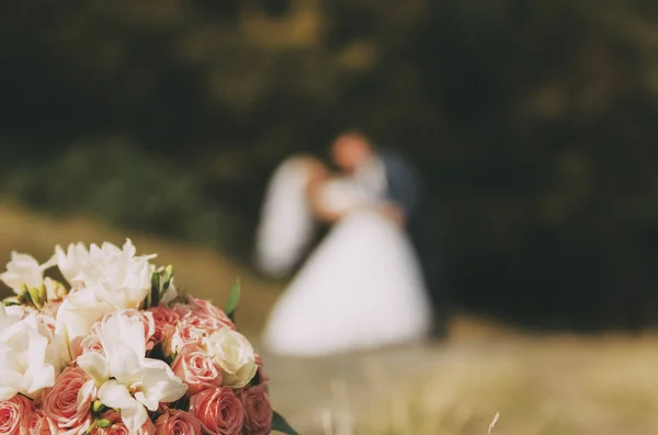 Buquê de casamento que coloca na grama com casal de casamento borrado — Fotografia de Stock