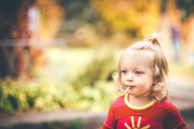 Sarışın ve mavi gözlü küçük şirin kız lolipopu yaladı, bulanık yaprakların arka planını.