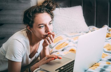 Serious young girl studying from home online during a quarantine virus outbreak. Solves tasks on a laptop in her room near the bed. Close-up. Stay at home concept. Solves issues with work. Copy space clipart