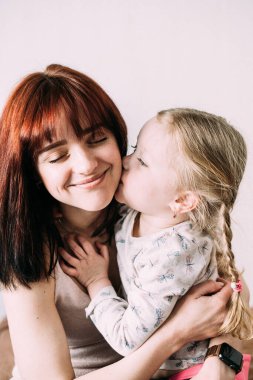 Sarışın bir çocuk, esmer annesine nazikçe sarılıyor ve yanağından öpüyor. Neşeli anne kapalı gözlerle gülümser. Hafif arkaplan. Anneler günü konsepti. Anne-kız ilişkisi.