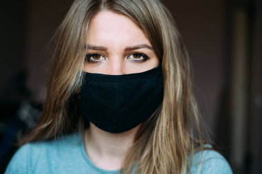 Siyah tıbbi maskeli, sarı saçlı, koyu renk gözlü ve turkuaz tişörtlü genç bir kadın kameraya bakıyor. Portre fotoğrafı. Karantinaya alındı. Evde kal konsepti. Salgın. Virüs. Koronavirüs. 