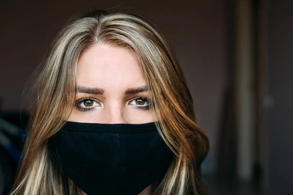 young woman in a medical mask with blond hair in a turquoise t-shirt is looking at the camera. portrait shooting. on a dark background. coronavirus. epidemic. virus. pandemic. stay home concept.