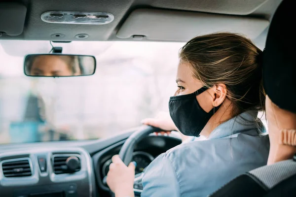 A young woman  taxi driver in a black medical mask with blond hair in a jacket rides a left-hand drive car. Woman do not look at camera. Stay home concept. Quarantine. Virus. Coronavirus. Pandemic.