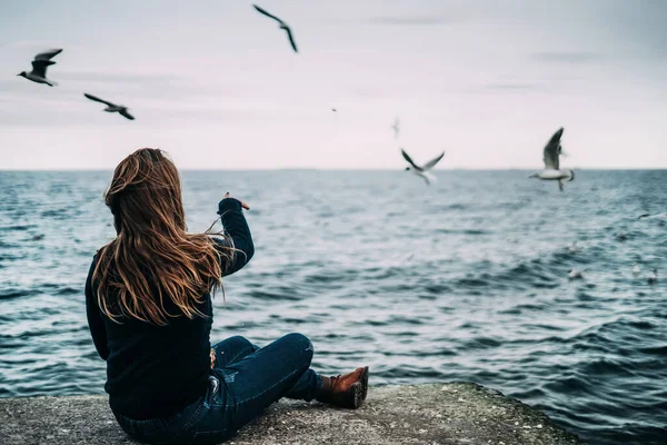Une jeune femme est assise dans un pull en tricot bleu et un jean au bord de la mer et nourrit les mouettes . — Photo